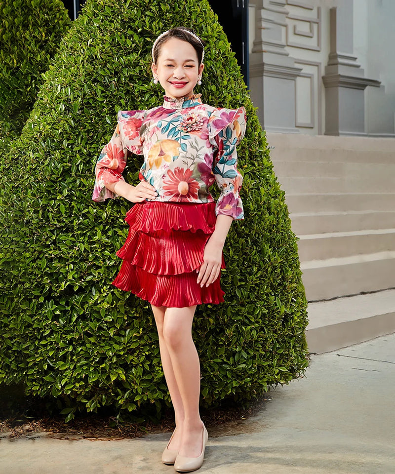 Woman in a floral top and red skirt standing next to a green bush