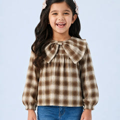 A young girl with long dark hair and pink hair clips is smiling, wearing a brown and beige plaid long-sleeve blouse with a large bow at the collar, paired with blue jeans, against a light blue background.