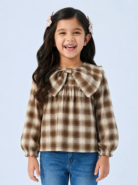 A young girl with long dark hair and pink hair clips is smiling, wearing a brown and beige plaid long-sleeve blouse with a large bow at the collar, paired with blue jeans, against a light blue background.