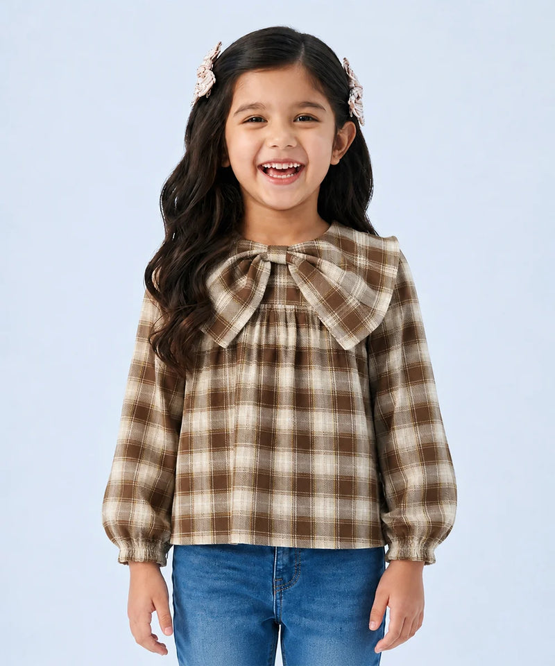 A young girl with long dark hair and pink hair clips is smiling, wearing a brown and beige plaid long-sleeve blouse with a large bow at the collar, paired with blue jeans, against a light blue background.