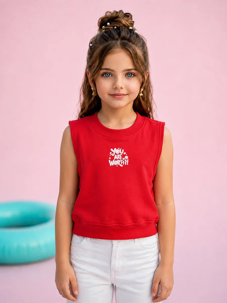 A young girl with her hair in a high bun stands against a solid pink background, wearing a bright red sleeveless crop top sweatshirt with a white graphic that reads 'Moms Just Want Off' and white pants.
