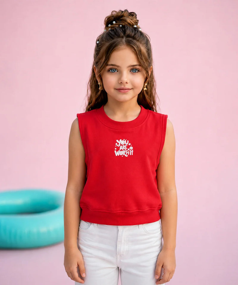 A young girl with her hair in a high bun stands against a solid pink background, wearing a bright red sleeveless crop top sweatshirt with a white graphic that reads 'Moms Just Want Off' and white pants.