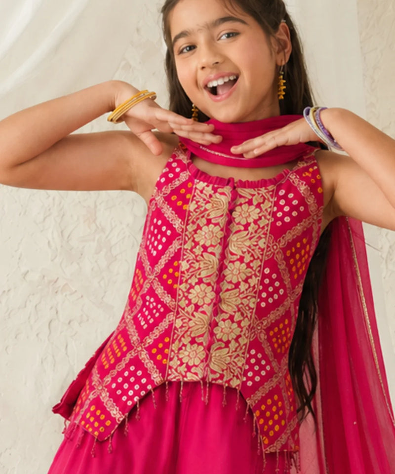 Young girl wearing a pink and gold traditional outfit with a scarf, smiling against a light background.