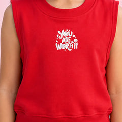 A close-up of a child wearing a bright red sleeveless sweatshirt with a white graphic text print that reads 'YOU ARE WORTH IT' on the chest, set against a solid light pink background.