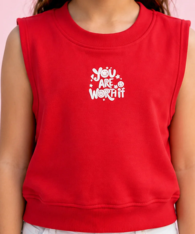 A close-up of a child wearing a bright red sleeveless sweatshirt with a white graphic text print that reads 'YOU ARE WORTH IT' on the chest, set against a solid light pink background.