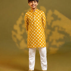 A child viewed from behind wearing a mustard yellow kurta with a white geometric diamond pattern, paired with white trousers and sandals, standing against a warm yellow background with soft shadow details.