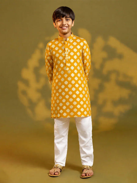 A child viewed from behind wearing a mustard yellow kurta with a white geometric diamond pattern, paired with white trousers and sandals, standing against a warm yellow background with soft shadow details.