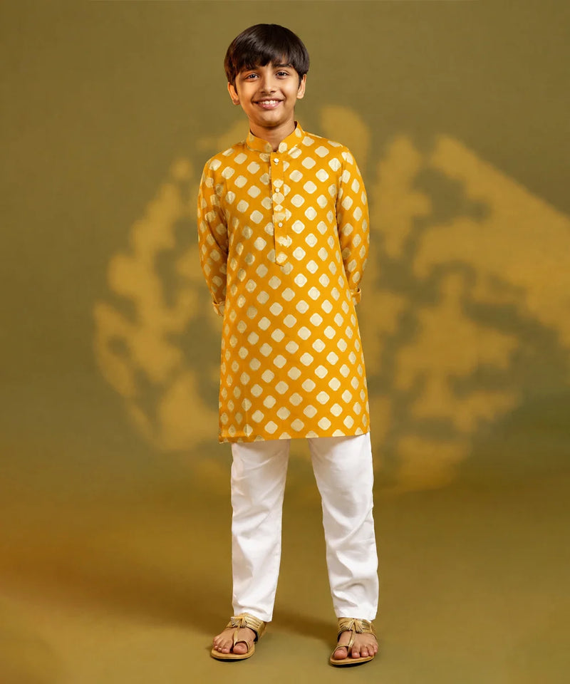 A child viewed from behind wearing a mustard yellow kurta with a white geometric diamond pattern, paired with white trousers and sandals, standing against a warm yellow background with soft shadow details.