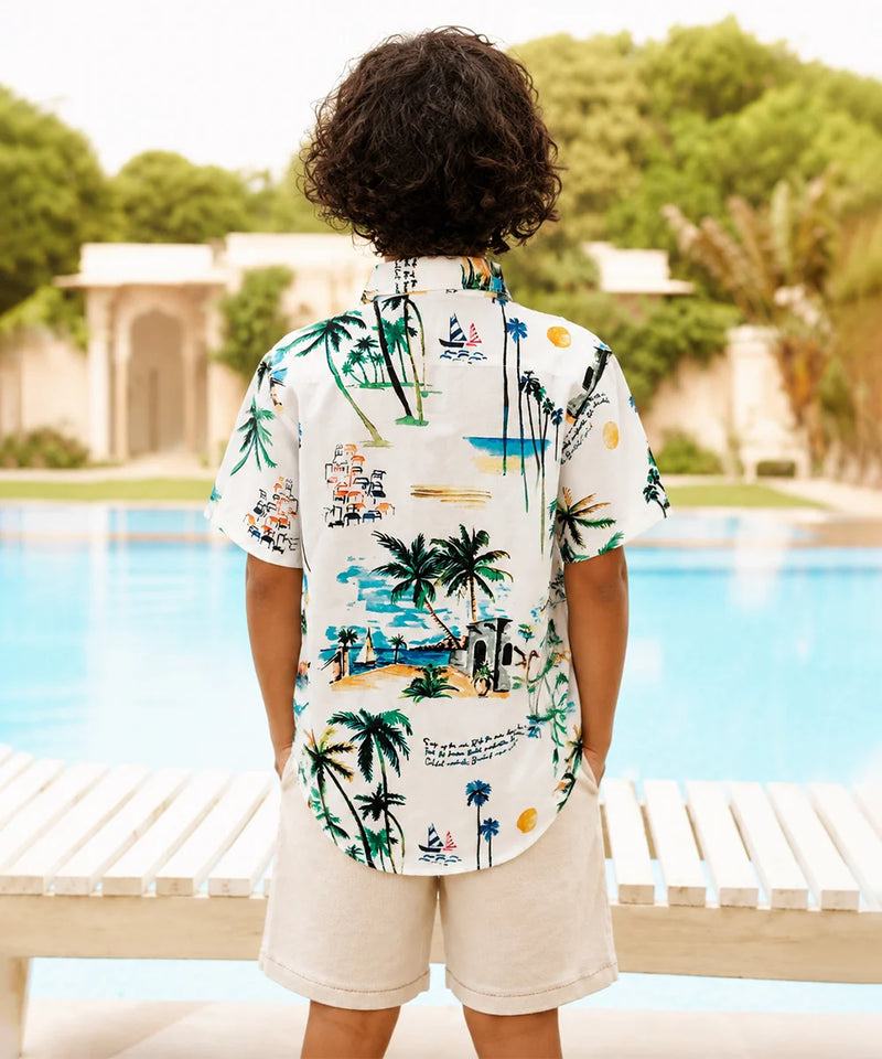 A child viewed from behind wearing a white short-sleeve button-up shirt with a colorful tropical beach print, standing next to a swimming pool.