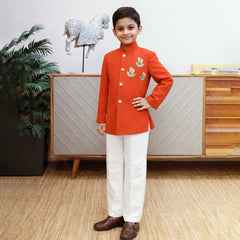 Young boy in a red traditional outfit standing in a room with a decorative cabinet and plant.