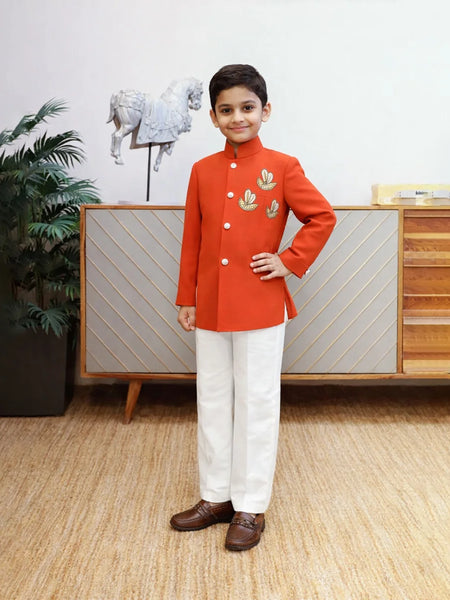 Young boy in a red traditional outfit standing in a room with a decorative cabinet and plant.