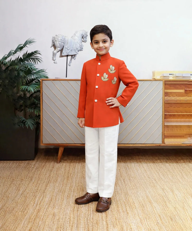 Young boy in a red traditional outfit standing in a room with a decorative cabinet and plant.