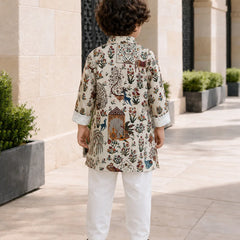 A child viewed from behind wearing a cream-colored kurta with an intricate multicolor floral and bird print, paired with white trousers and black sandals, standing on a stone walkway.