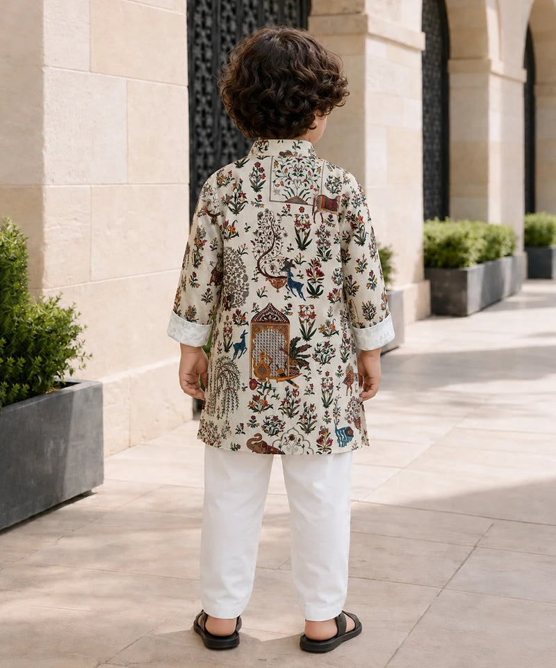 A child viewed from behind wearing a cream-colored kurta with an intricate multicolor floral and bird print, paired with white trousers and black sandals, standing on a stone walkway.