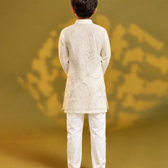 A child viewed from behind wearing a white embroidered kurta and white trousers, standing against a warm olive-gold studio background with soft shadow patterns.