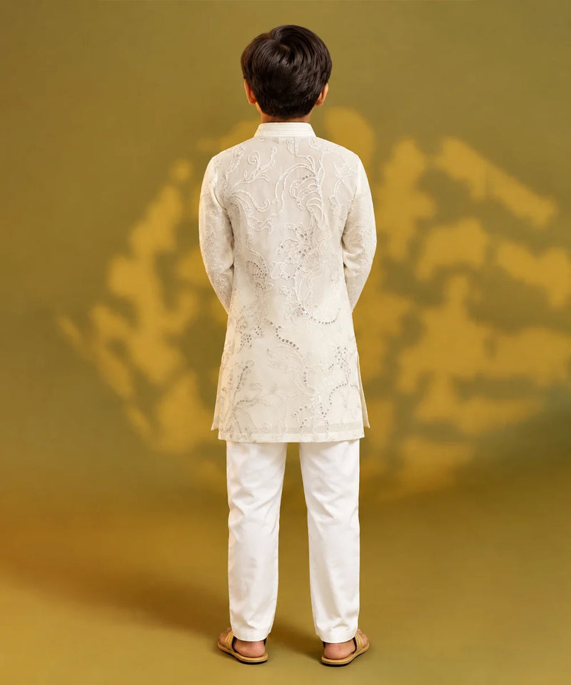 A child viewed from behind wearing a white embroidered kurta and white trousers, standing against a warm olive-gold studio background with soft shadow patterns.