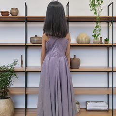 Girl in a long purple dress standing in front of a bookshelf with decorative items.