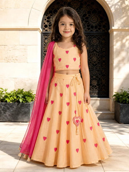 Young girl in a peach dress with red heart patterns and a pink dupatta standing in front of an archway.