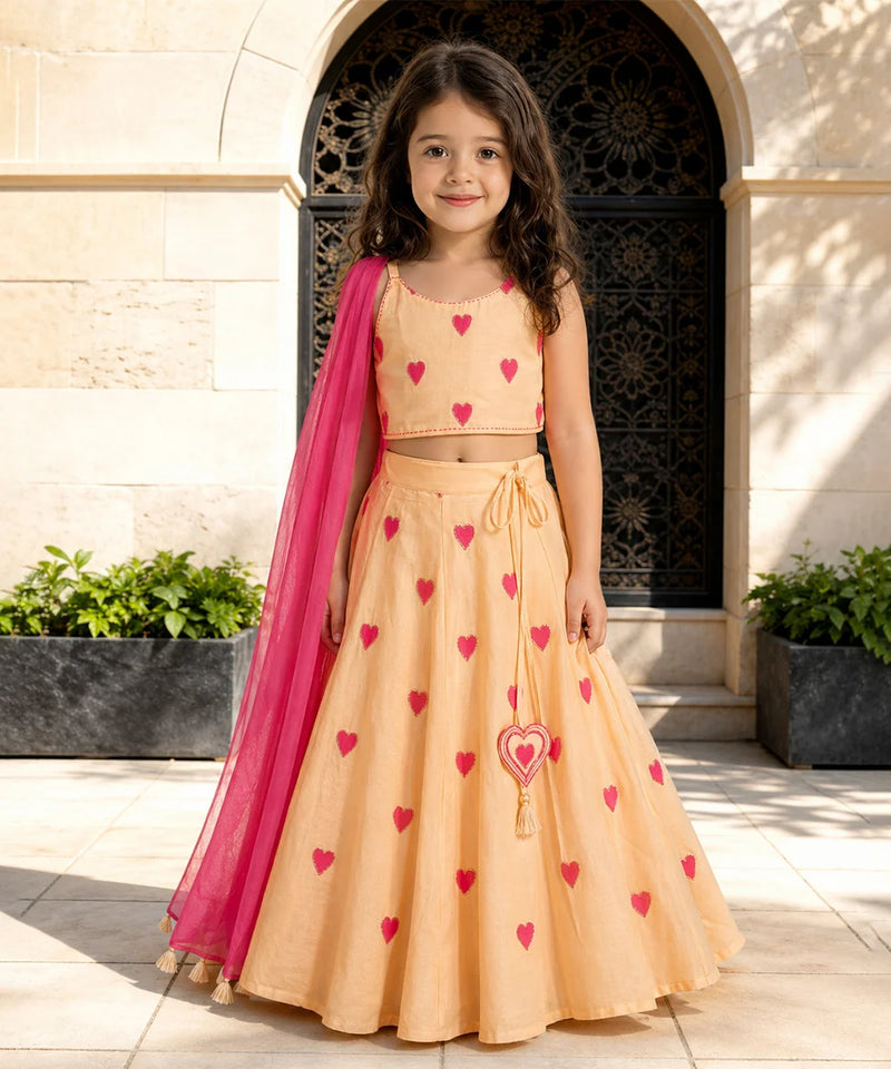 Young girl in a peach dress with red heart patterns and a pink dupatta standing in front of an archway.