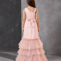 A young girl viewed from the back wearing a light pink formal dress with a one-shoulder design, a large bow at the waist, and a tiered tulle skirt, standing against a textured gray studio backdrop.