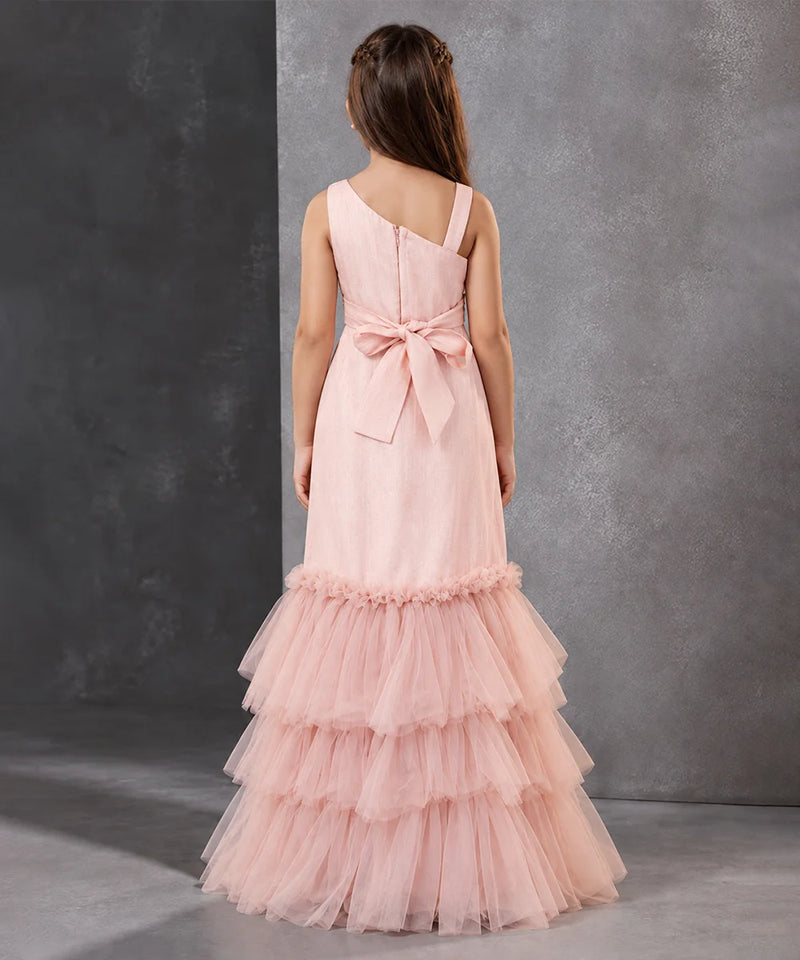 A young girl viewed from the back wearing a light pink formal dress with a one-shoulder design, a large bow at the waist, and a tiered tulle skirt, standing against a textured gray studio backdrop.