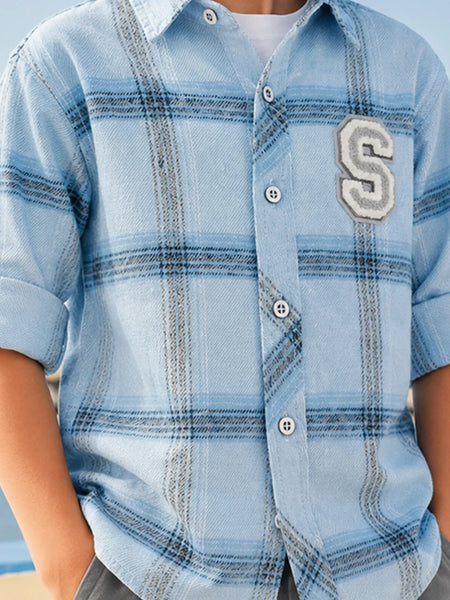 A close-up of a child wearing a light blue plaid short-sleeve button-up shirt with a large gray and white embroidered letter 'S' patch on the left chest, over a white undershirt.