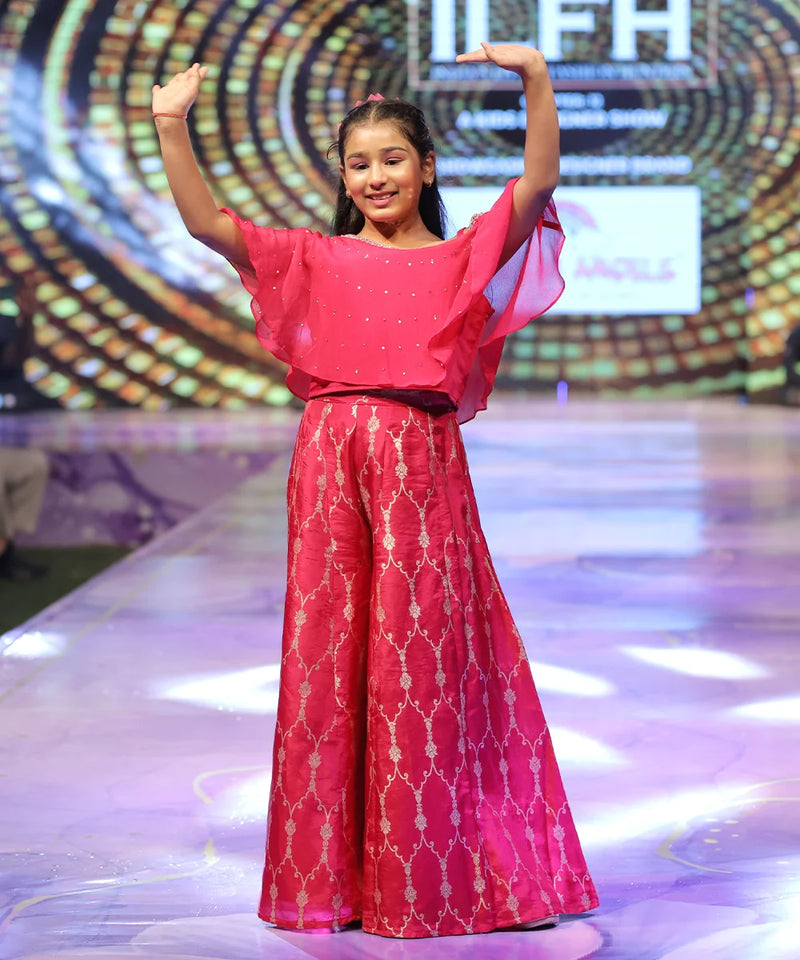 Young girl in a red sharara outfit with a patterned design on a stage with a colorful background