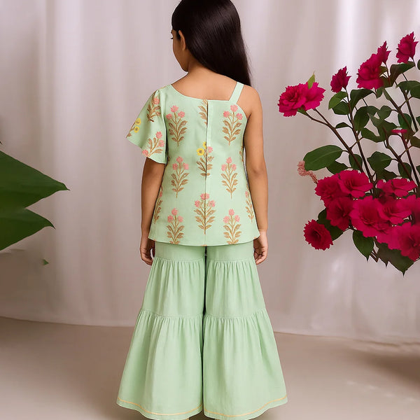 Woman wearing a light green outfit with floral patterns, standing against a white curtain with pink flowers.