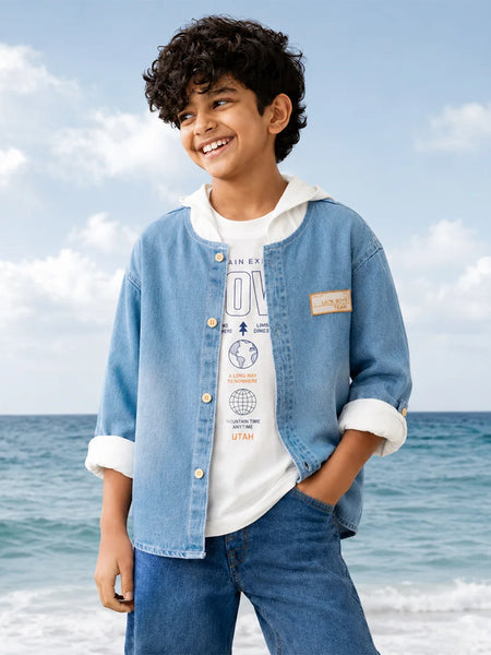 A smiling boy standing on a beach wearing a light blue denim jacket over a white graphic T-shirt and blue jeans, with ocean and sky in the background.