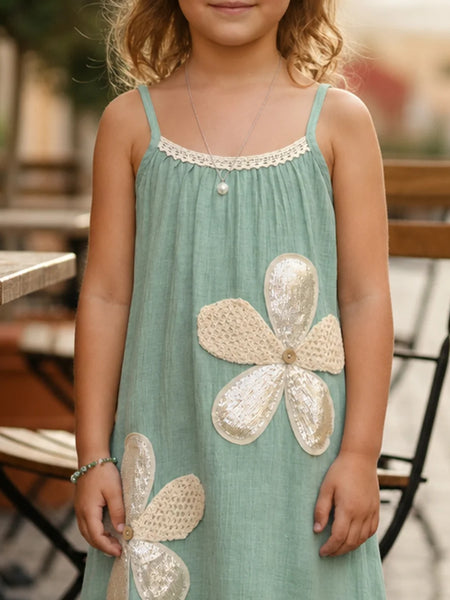 A young girl wearing a sage green sleeveless dress with large textured cream flower appliqués and a lace-trimmed neckline, photographed in an outdoor patio setting.