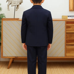 Person wearing a navy suit standing in front of a wooden cabinet with decorative items.