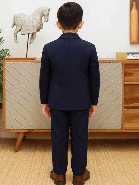 Person wearing a navy suit standing in front of a wooden cabinet with decorative items.