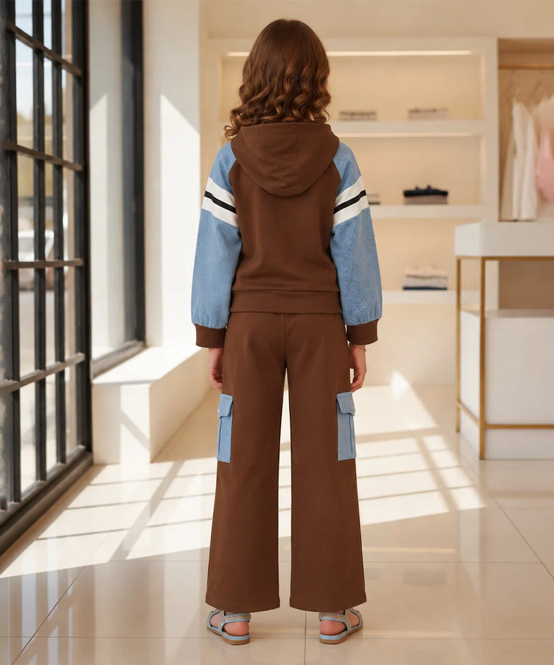 A child model viewed from behind wearing a brown hooded sweatshirt and matching wide-leg pants with light blue color-blocked sleeve and pocket panels, standing in a bright indoor boutique setting.