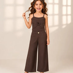 A young girl with long wavy brown hair wearing a dark brown two-piece outfit consisting of a sleeveless top with a ruffled flower detail and matching wide-leg pants, paired with light blue shoes, standing against a warm beige background with soft window pane shadows.