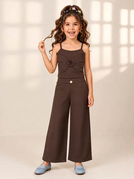A young girl with long wavy brown hair wearing a dark brown two-piece outfit consisting of a sleeveless top with a ruffled flower detail and matching wide-leg pants, paired with light blue shoes, standing against a warm beige background with soft window pane shadows.