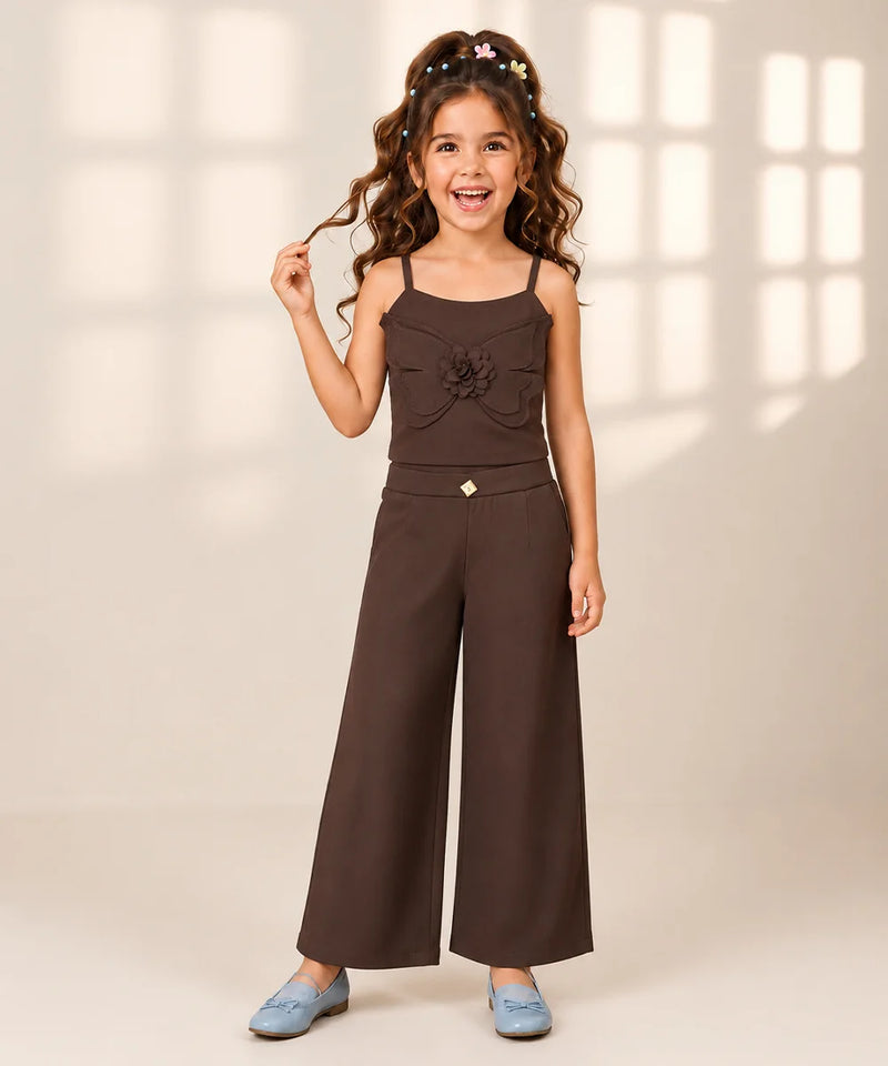 A young girl with long wavy brown hair wearing a dark brown two-piece outfit consisting of a sleeveless top with a ruffled flower detail and matching wide-leg pants, paired with light blue shoes, standing against a warm beige background with soft window pane shadows.