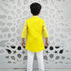 Child wearing a yellow kurta with white pants standing against a decorative wall.