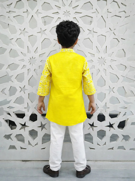 Child wearing a yellow kurta with white pants standing against a decorative wall.