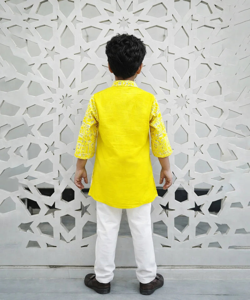 Child wearing a yellow kurta with white pants standing against a decorative wall.