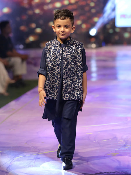 Child model walking on a fashion show runway wearing a navy blue outfit with a patterned vest.