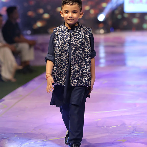 Child model walking on a fashion show runway wearing a navy blue outfit with a patterned vest.