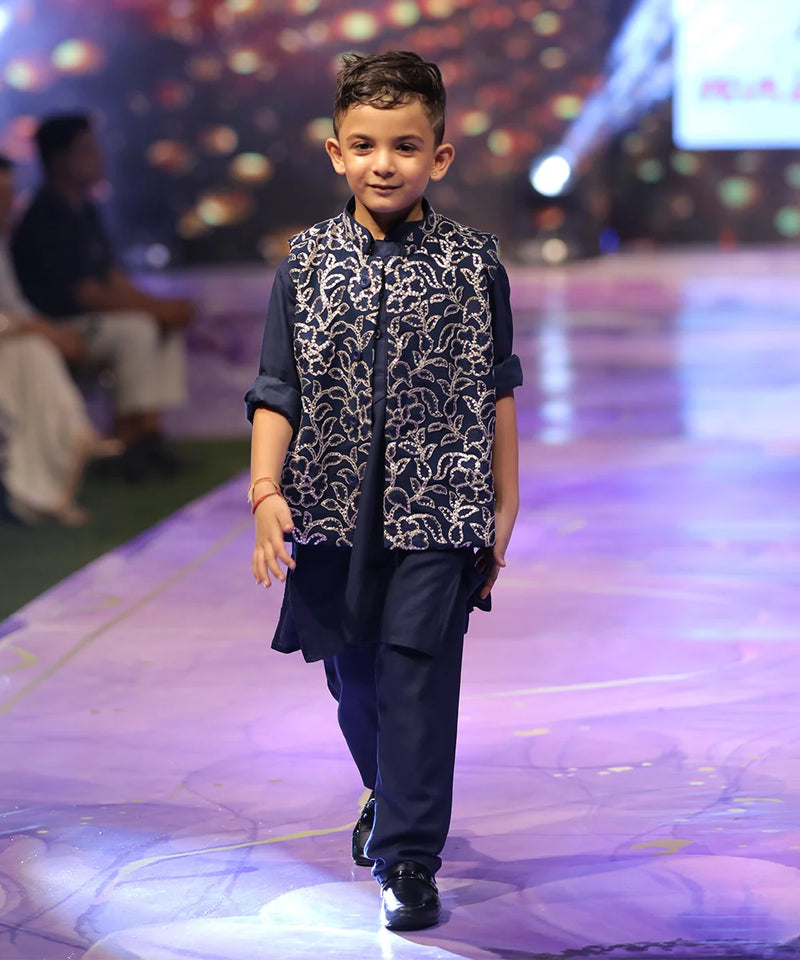 Child model walking on a fashion show runway wearing a navy blue outfit with a patterned vest.