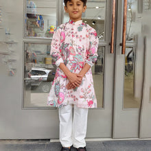 Child wearing a floral kurta with white pants standing in front of a glass door.