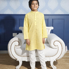 Child wearing a yellow kurta with white pants standing in front of a decorative chair.