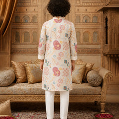 A person standing with their back to the camera, wearing a cream-colored kurta with pink, green, and blue geometric and floral patterns, paired with white trousers, in an ornate traditional room with gold-toned furniture.