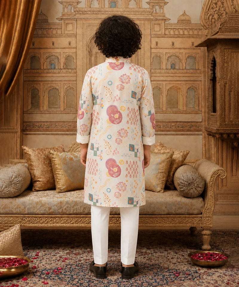 A person standing with their back to the camera, wearing a cream-colored kurta with pink, green, and blue geometric and floral patterns, paired with white trousers, in an ornate traditional room with gold-toned furniture.