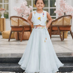 Young girl in a light blue traditional outfit standing on steps with decorative flowers in the background.