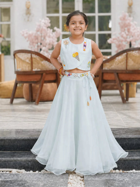 Young girl in a light blue traditional outfit standing on steps with decorative flowers in the background.