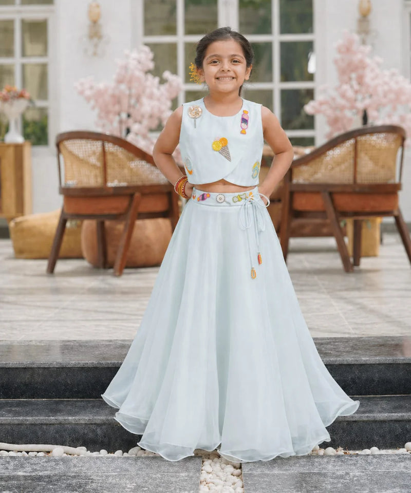 Young girl in a light blue traditional outfit standing on steps with decorative flowers in the background.