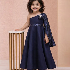Young girl in a navy blue one-shoulder dress with sequin sleeves standing next to a small table against a pink background.